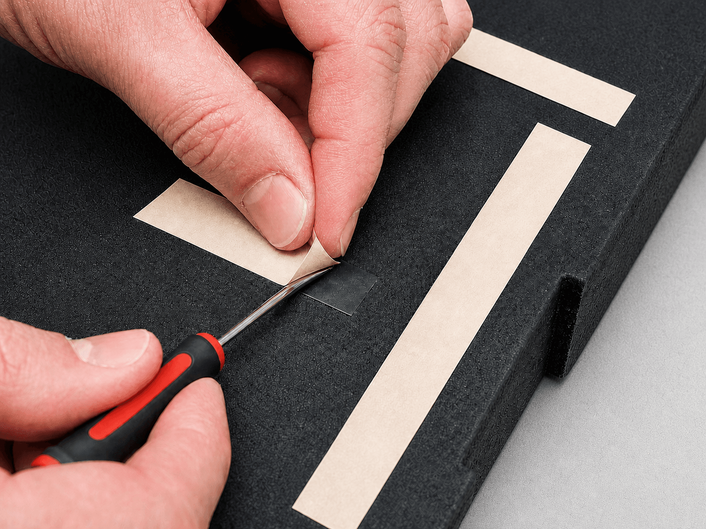 A small flat-head screwdriver gently lifting the corner of an adhesive strip's paper backing on a black foam insert