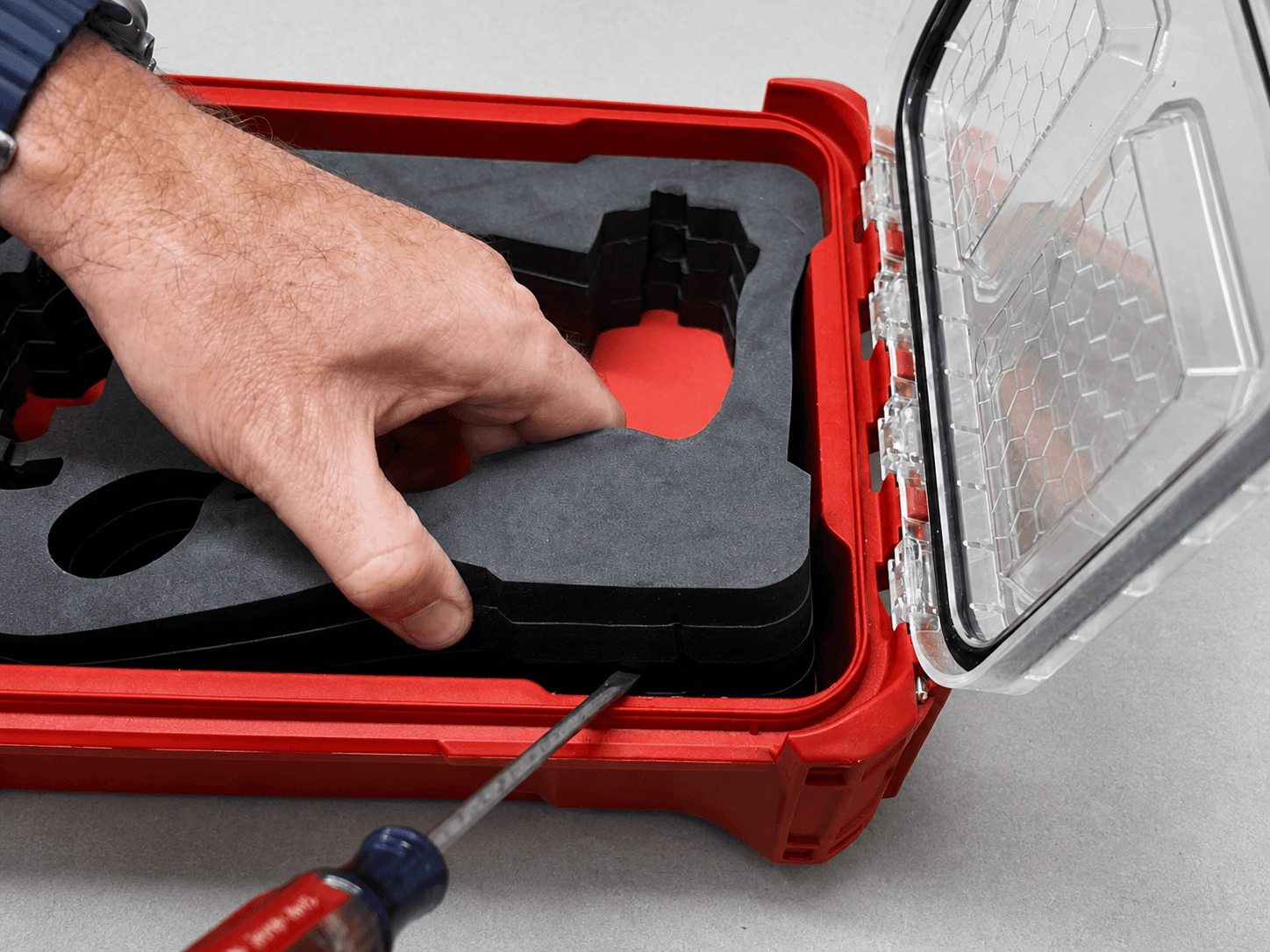 A flat-head screwdriver gently starting a gap between a black foam insert and the wall of a red hard case while the insert is lifted by hand